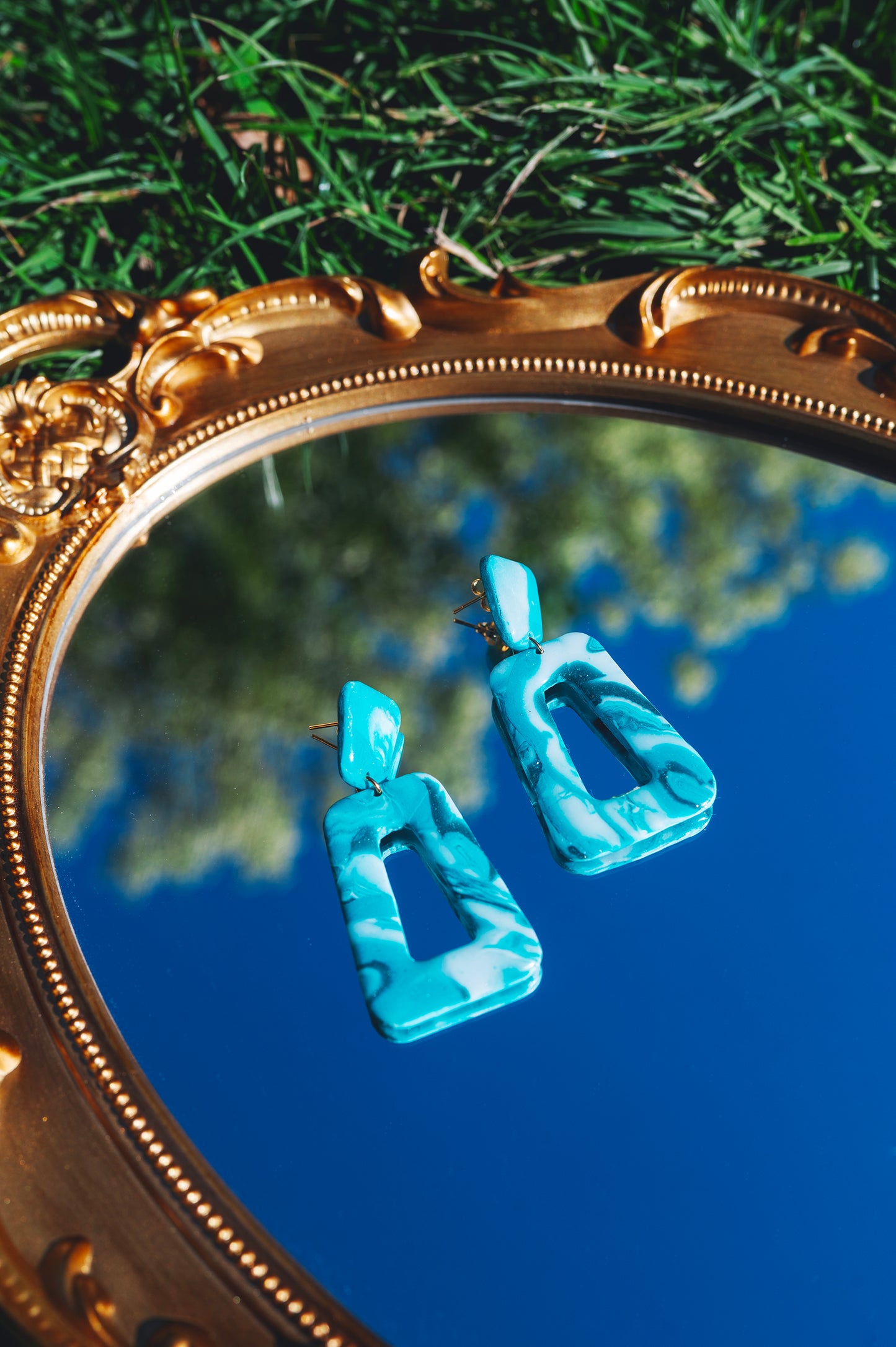 deux boucles d'oreilles fantaisie en argile polymère faites à la main, constituées d'un petit trapèze et d'un grand trapèze turquoises, posés sur un miroir qui reflète le ciel bleu