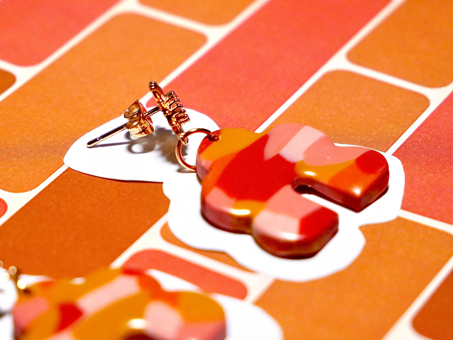 une boucle d'oreilles rose, ocre et rougefaites à la main en argile polymère, en forme d'arche et de nuage, avec un apprêt en acier inoxydable plaqué or en forme de soleil avec un zicron.