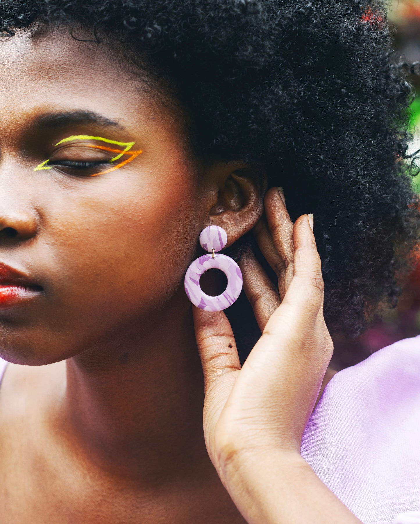 visage de femme noire de profil avec un maquillage coloré, tenant entre ses doigt la boucle d'oreille fantaisie en argile polymère faites main qu'elle porte, composé d'un petit rond au dessus d'un grand cercle