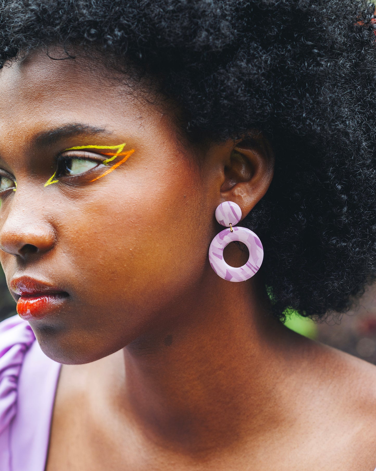 visage de femme noire de profil avec un maquillage coloré, portant une boucle d'oreille fantaisie en argile polymère faites au dessus d'un grand cercle