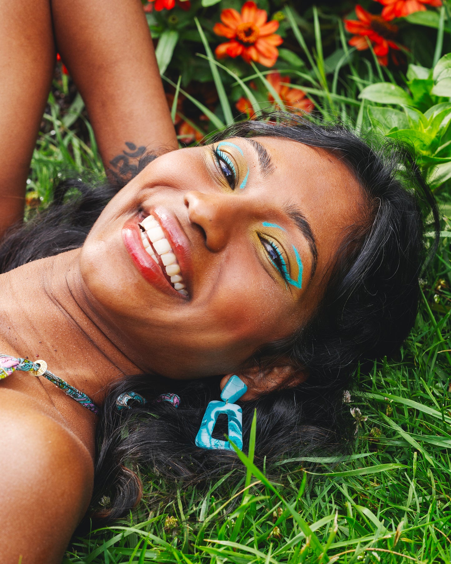 visage de femme indienne allongée dans l'herbe, portant deux boucles d'oreilles fantaisie en argile polymère faites à la main, constituées d'un petit trapèze et d'un grand trapèze turquoises