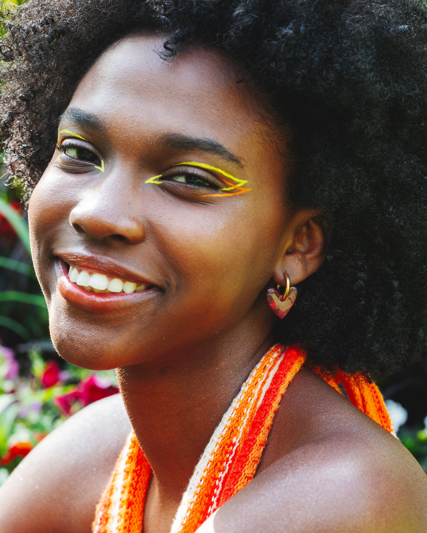 femme à la peau noire souriante au maquillage eye-liner coloré, portant deux boucles d'oreilles fantaisie en forme de coeurs rouges, fait main en argile polymère, avec des anneaux doré en acier inoxydable