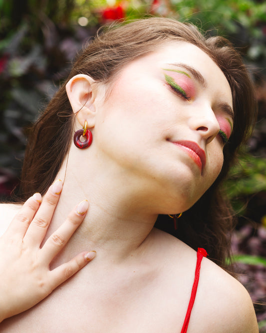 visage de femme aux yeux fermés portant un maquillage coloré vert et rose et des boucles d'oreilles fantaisie faites à la main en argile polymère rouge, avec un anneau doré en acier inoxydable