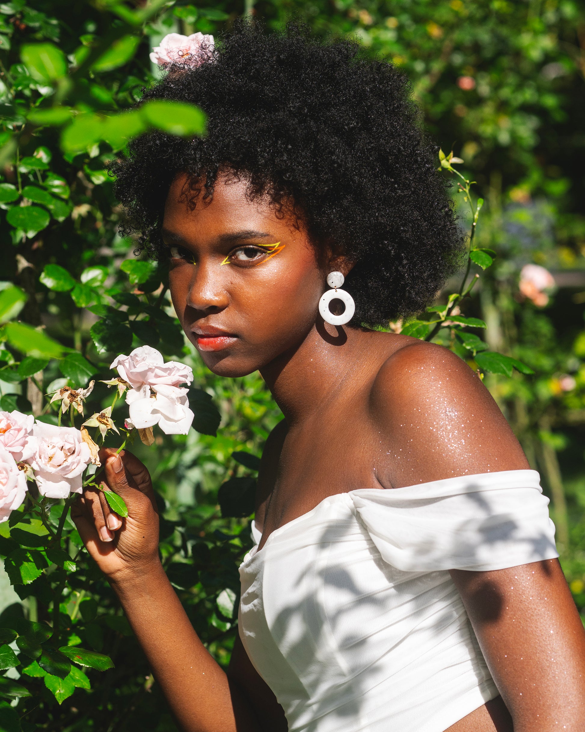 femme noire dans un jardin tenant une rose, au maquillage coloré orange et jaune, portant une boucle d'oreille fantaisie faite main en argile polymère, composée d'un petit rond et d'un grand cercle, de couleur blanche avec des points noirs incrusté, et un anneau doré en acier inoxydable