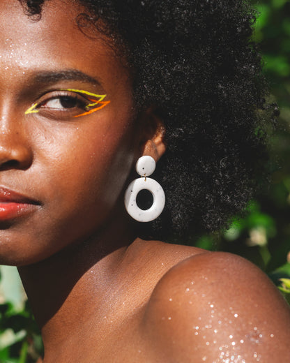 visage d'une femme noire au maquillage coloré orange et jaune, portant une boucle d'oreille fantaisie faite main en argile polymère, composée d'un petit rond et d'un grand cercle, de couleur blanche avec des points noirs incrusté, et un anneau doré en acier inoxydable