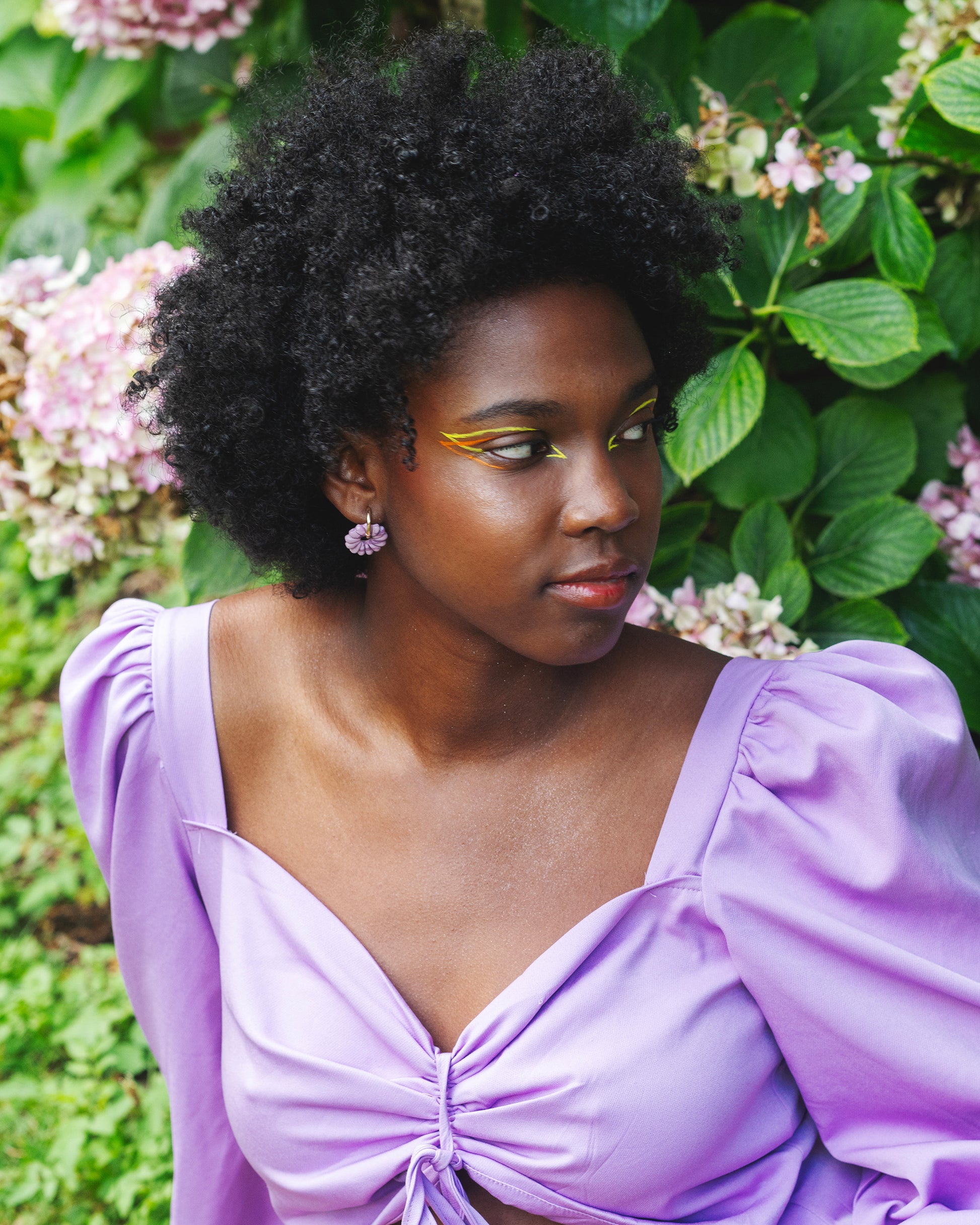 femme noire assise dans une jardin fleuri portant un maquillage au liners jaune et orange et une paire de boucles d'oreilles fantaisies faites à la main, violette et de forme de fleur de marguerite, montée sur un anneau rond doré en acier inoxydable