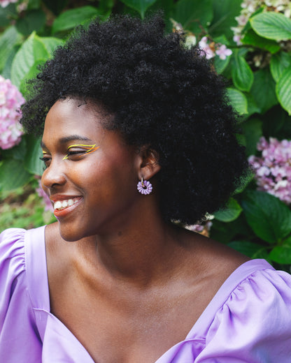 visage de femme noire souriant, portant un maquillage au liners jaune et orange et une paire de boucles d'oreilles fantaisies faites à la main, violette et de forme de fleur de marguerite, montée sur un anneau rond doré en acier inoxydable