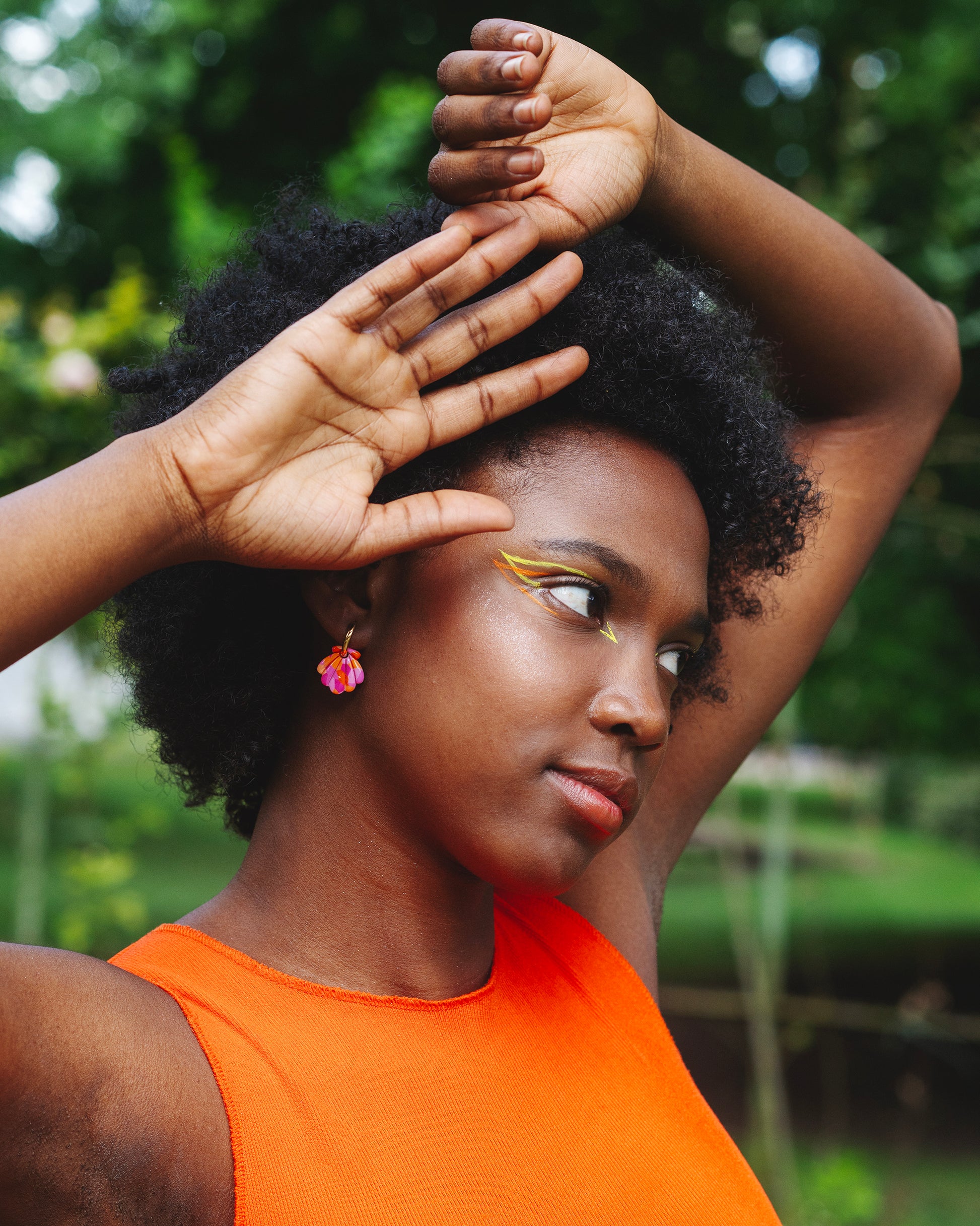 femme noire de profil dans un jardin au maquillage liner jaune et orange, robe orange, qui porte une boucle d'oreille fantaisie en forme de coquillage en argile polymère et anneau doré en acier inoxydable