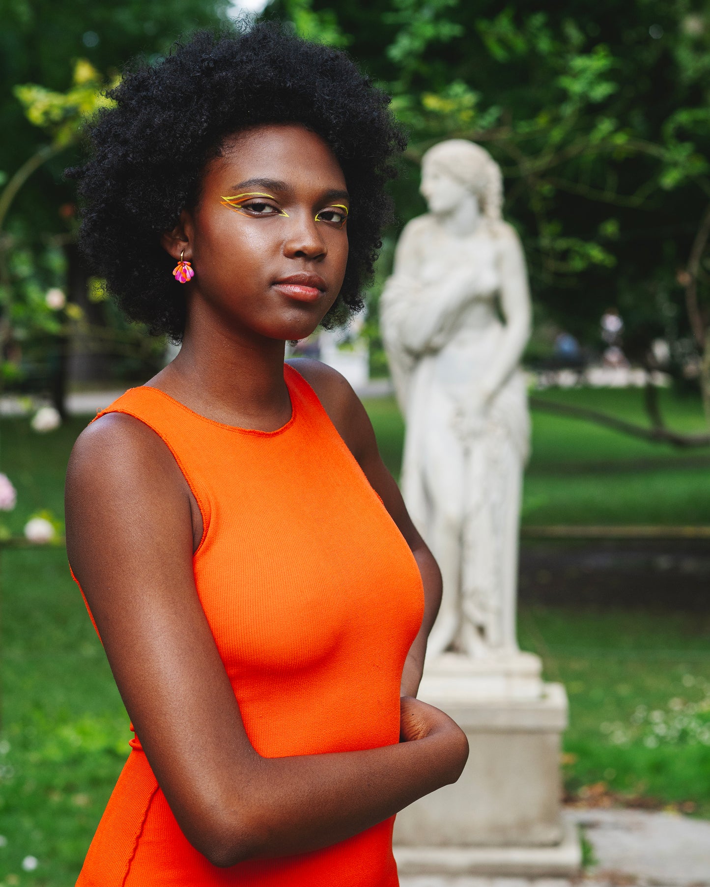 femme noire avec une robe orange et de l'eye-liner orange et jaune portant une boucle d'oreille fantaisie en forme de coquillage orange et rose en argile polymère sur un anneau doré en acier inoxydable dans un jardin avec une statue de style antique en fond