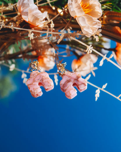 deux boucles d'oreilles fantaisie en forme de nuage et d'archeen argile polymère rose quartz faites à la main, avec un apprêt plaqué or 18 carraats en forme de soleil, posées sur un miroir reflétant le ciel bleu