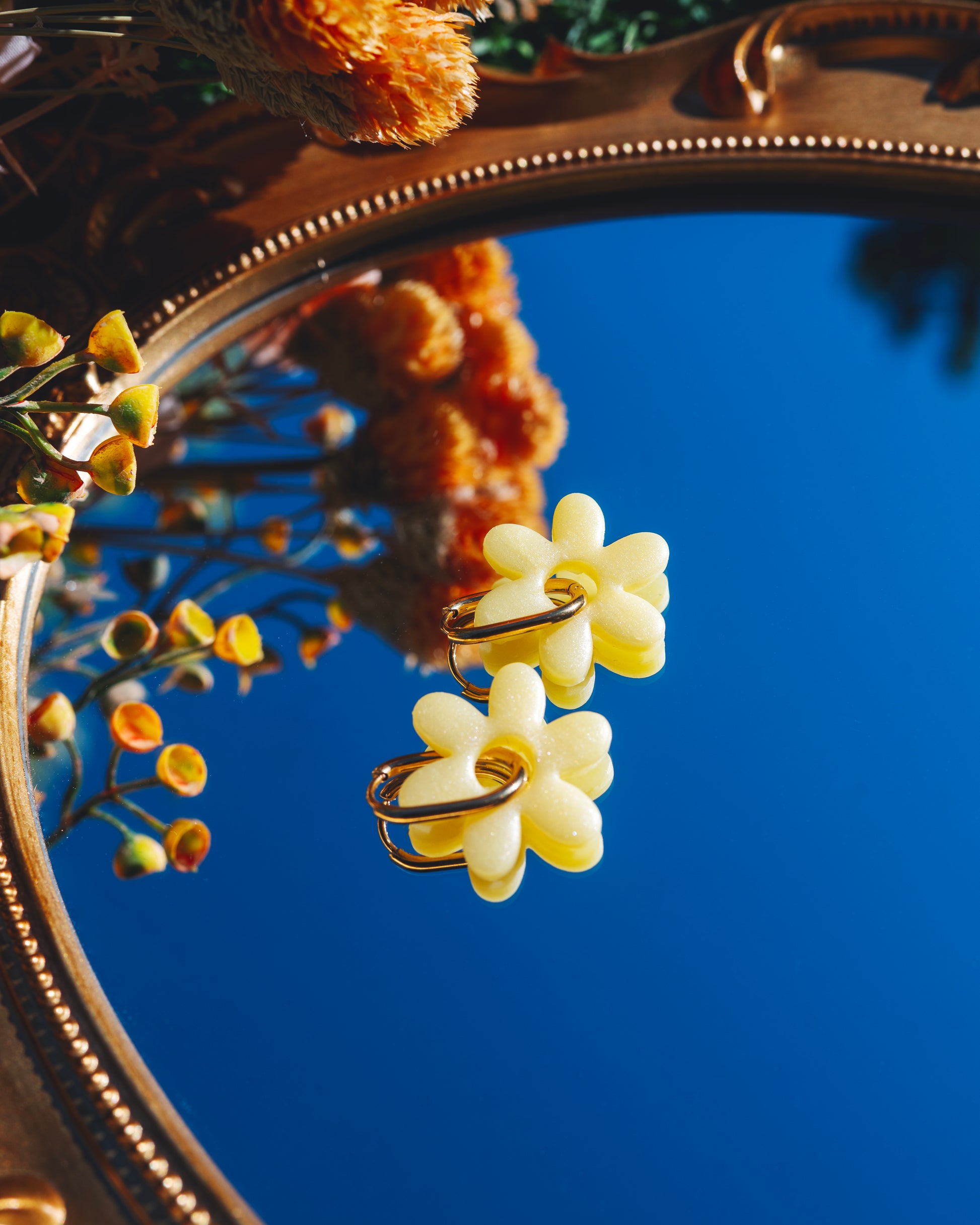 deux boucles d'oreilles jaunes en forme de fleur de jasmin en argile polymère faites à la main, avec deux anneaux doré en acier inoxydable, posées sur un miroir reflétant le ciel bleu