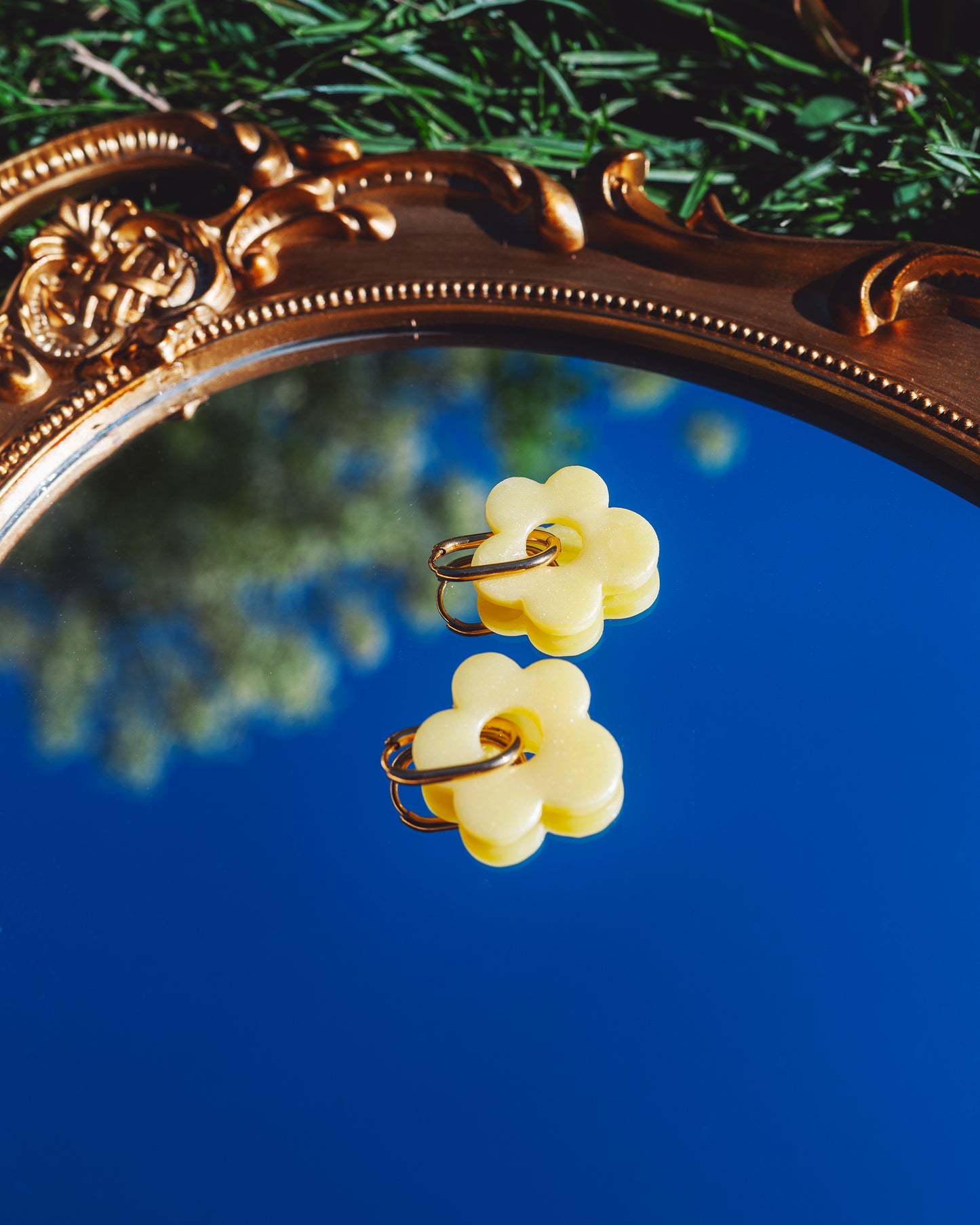 deux boucles d'oreilles fantaisie faite main en forme de fleur jaune, avec un anneau en acier inoxydable, posées sur un miroir reflétant le ciel bleu