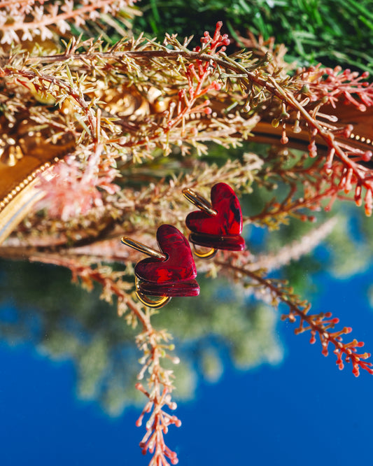 deux boucles d'oreilles fantaisie en forme de coeurs rouges, fait main en argile polymère, avec des anneaux doré en acier inoxydable, posés sur un miroir reflétant le ciel