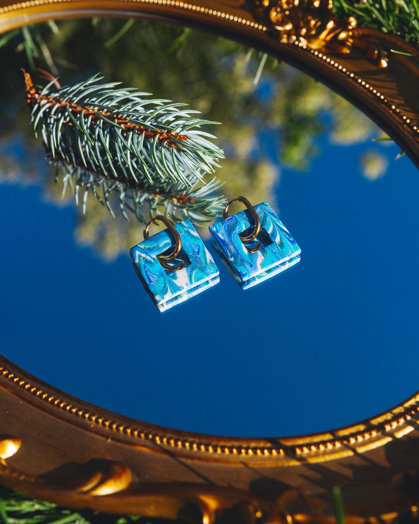 deux boucles d'oreilles fantaisie en argile polymère bleues avec anneaux rectangulaires dorés en acier inoxydable, posées sur un mirroir reflétant le ciel bleu