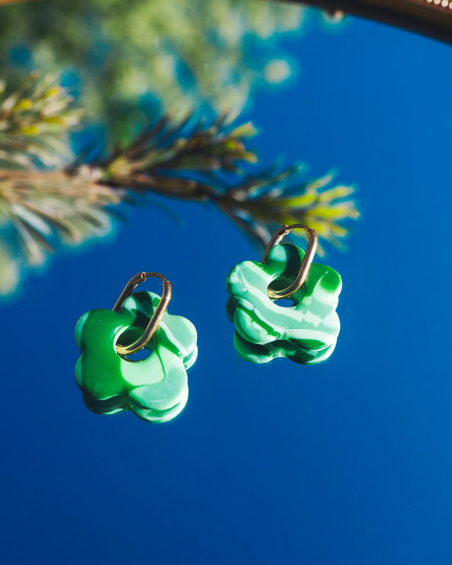 deux bocles d'oreilles en argile polymère faites main en forme de fleur, de couleurs vertes, avec des anneaux rectangulaire dorés en acier inoxydable, sur un miroir reflétant le ciel bleu