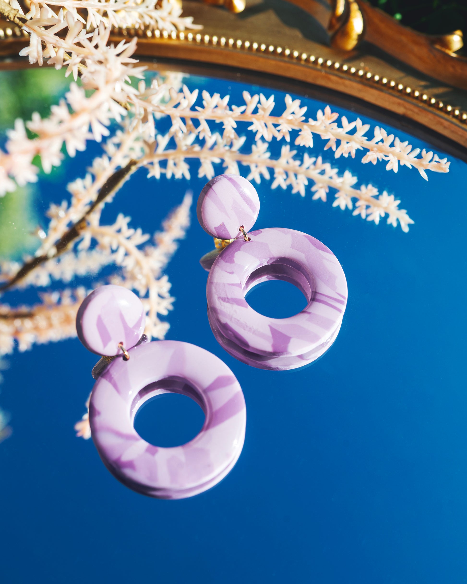 deux boucles d'oreilles en argile polymère faites main, violettes marbrées, composées d'un petit rond au dessus d'un grand cercle, posées sur un miroir reflétant le ciel bleu