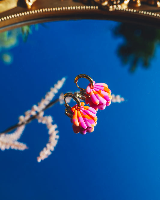 paire de boucles d'oreilles fantaisie en forme de coquillage en argile polymère rose et orange avec des anneaux dorés en acier inoxydable sur un mirroir qui  reflète le ciel bleu