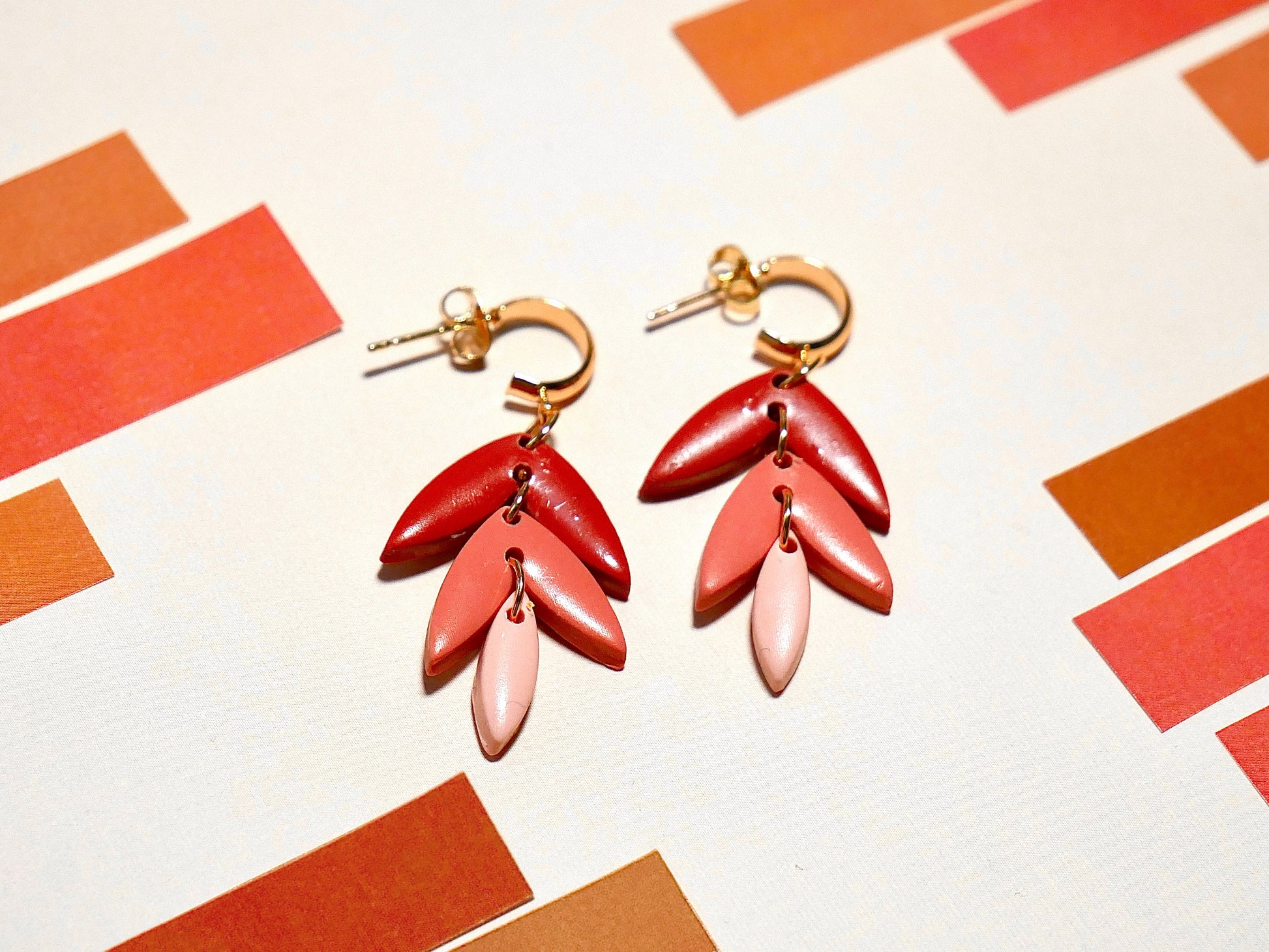 deux boucles d'oreilles fantaisie aux couleurs rouges et roses dégradées, et divisées en trois parties, le tout en forme de goutte, en argile polymère et faite main, sur fond blanc et brique