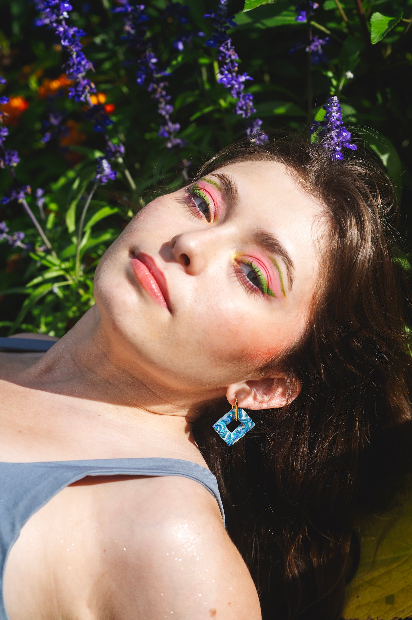 visage de femme allongée dans les fleurs au maquillage vert et rose, portant une boucle d'oreille fantaisie bleue en argile polymère sur un anneau rectangulaire doré en acier inoxydable