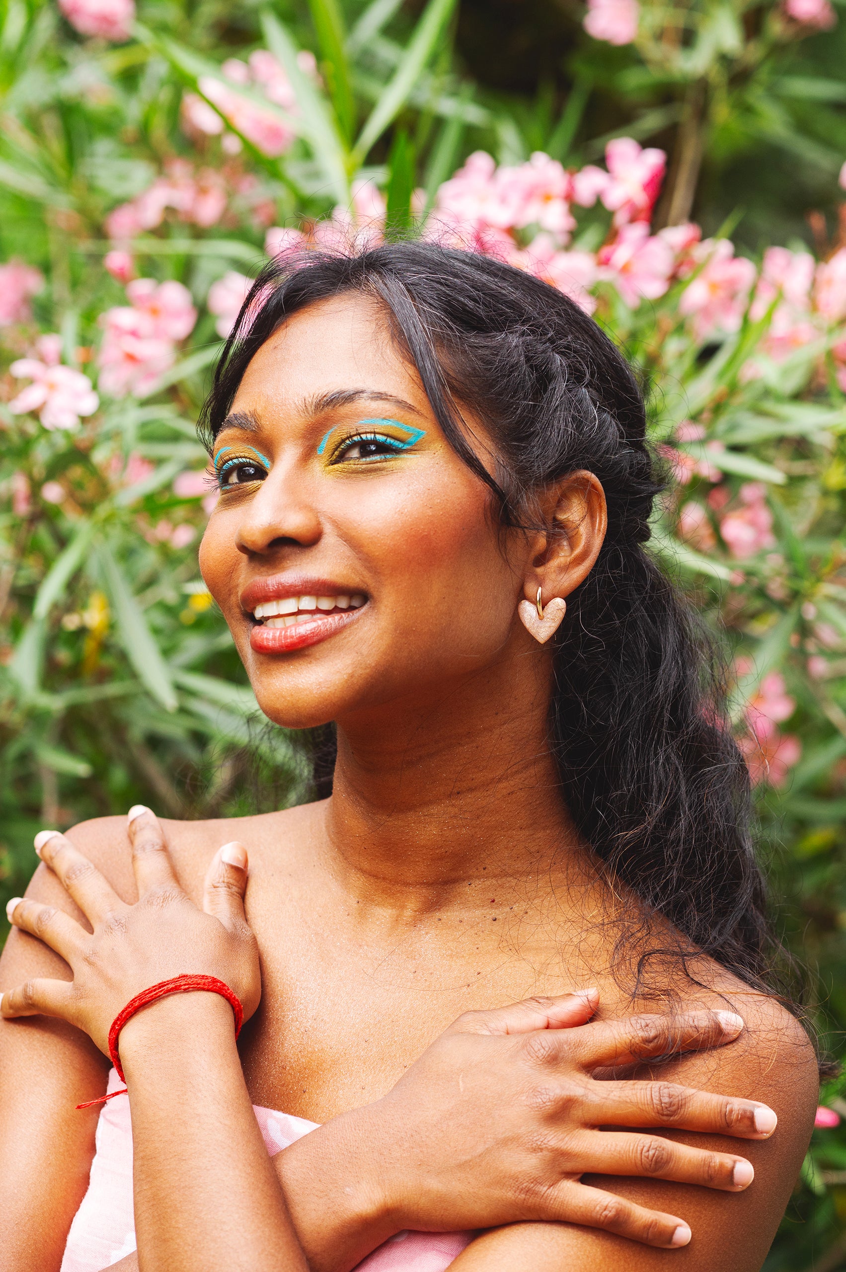 visage de femme indienne croisant les mains sur les épaule avec un maquillage coloré turquoise, posant devant des hortensias roses, et portant des boucles d'oreilles fantaisie roses quartz en forme de coeur, faites à la main en argile polymère