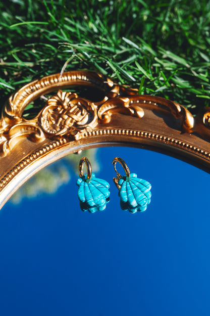 deux boucles d'oreilles fantaisie en forme de coquillages bleues turquoises avec des anneaux dorés en acier inoxydable, posées sur un miroir qui reflète le ciel bleu