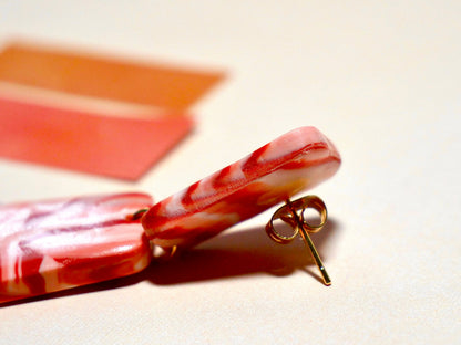 détail zoomé sur une boucle d'oreille en argile polymère faites à la main au motif de brique, aux couleurs rouge et blanc, constituées de deux trapèze, sur fond blanc et briques, avec détail sur l'attache en acier inoxydable doré