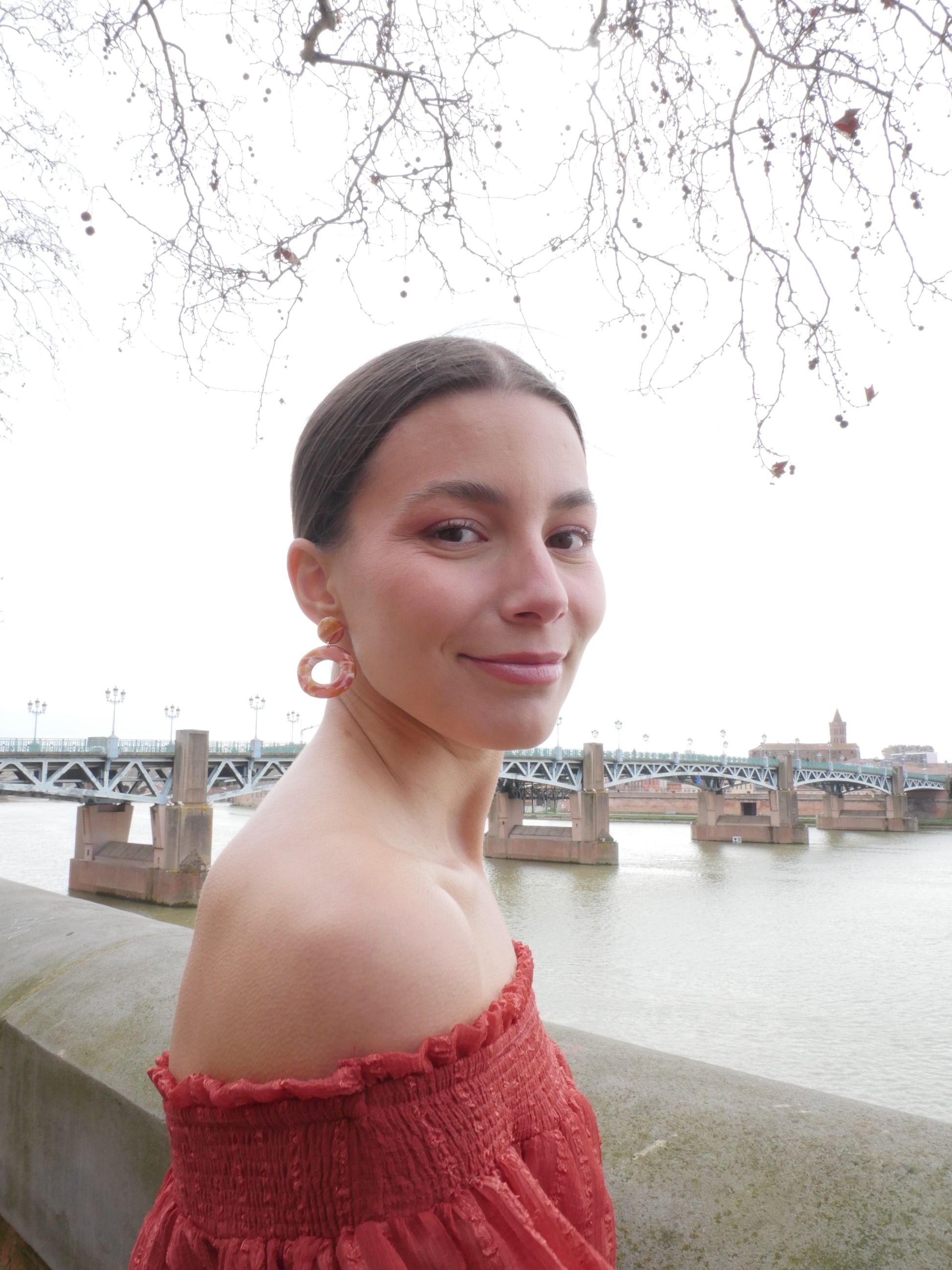 femme souriante de profil devant le pont Saint Pierre à Toulouse portant une boucle d'oreille composée d'un rond et un grand cercle rouges, roses et oranges