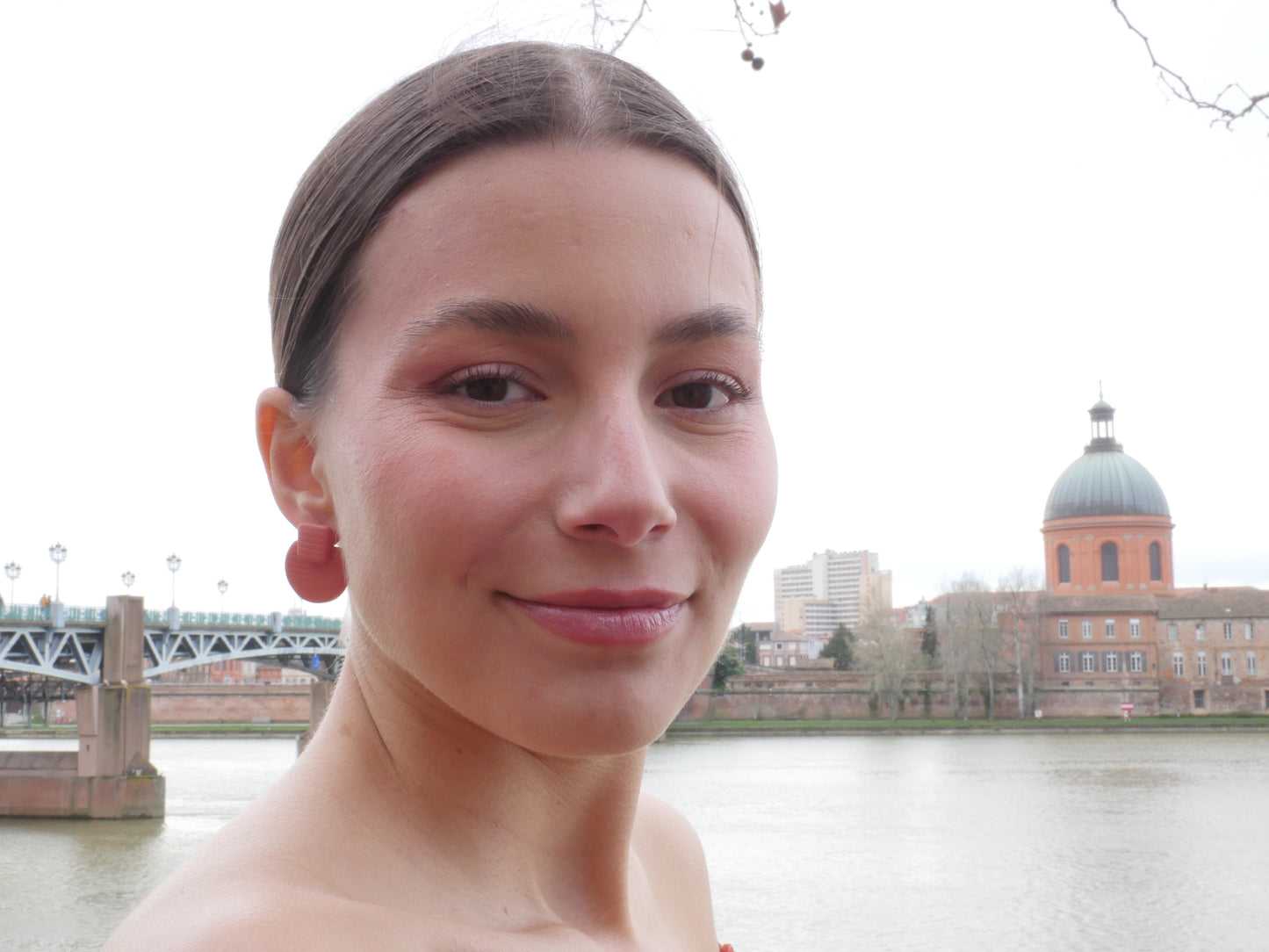 visage d'une femme de face  qui porte une boucle d'oreille en argile polymère rayée terracotta avec en fond le Dôme de la Grave à Toulouse