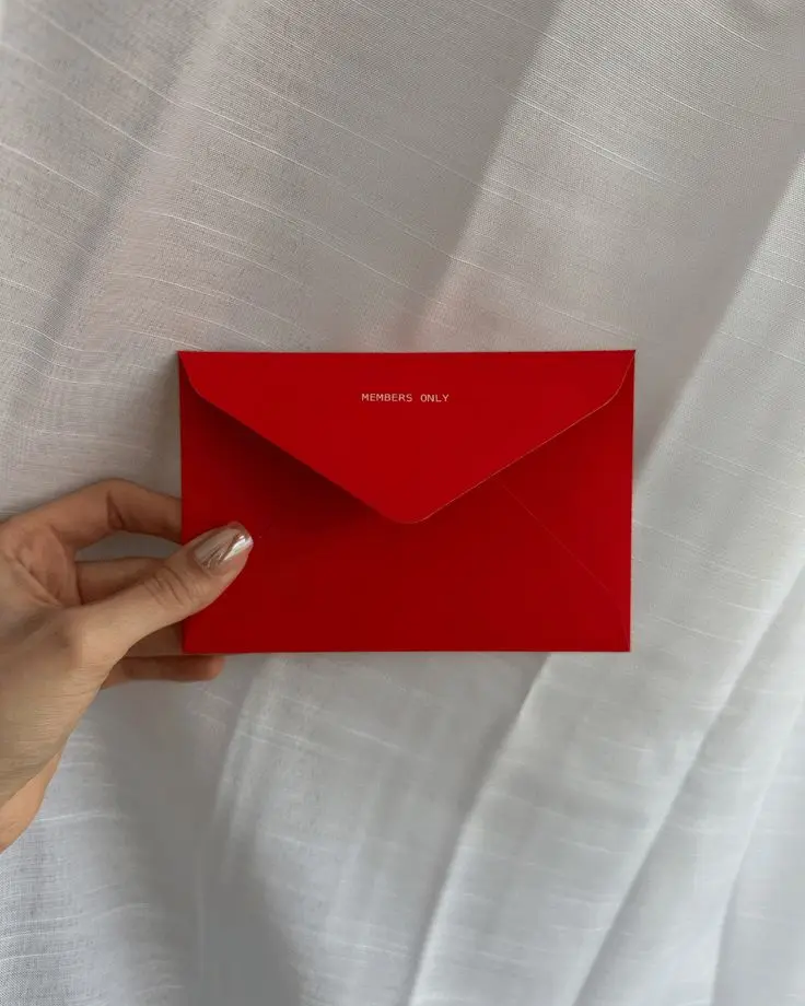 Main tenant une petite enveloppe rouge marquée « Members only » sur un drap blanc
