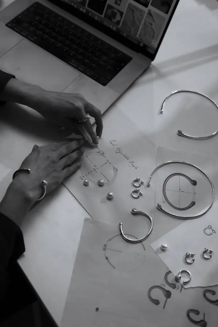 Main posée sur un croquis de bijou, entourée de joncs et de boucles d'oreilles en argent, en noir et blanc